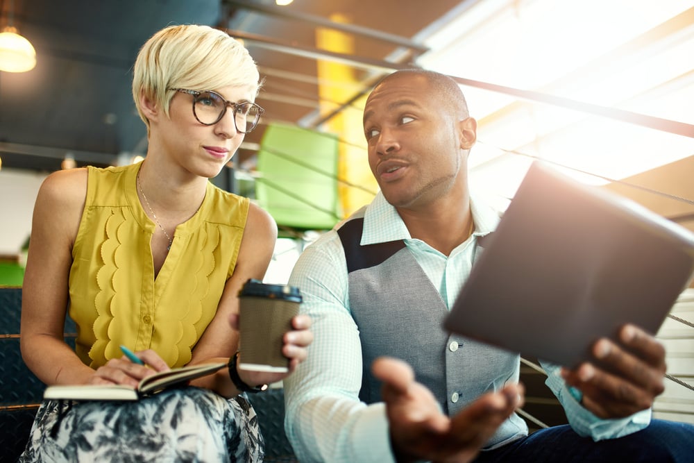 Two creative millenial small business owners working on social media strategy using a digital tablet while sitting in staircase Two creative millenial small business owners working on social media strategy using a digital tablet while sitting in staircase